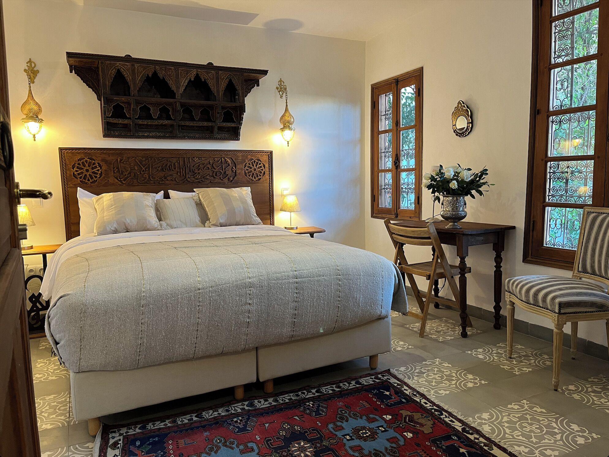 Un dormitorio con una cama grande, cabecera de madera, dos lámparas fijadas a la pared, una mesita con un florero y una silla.
