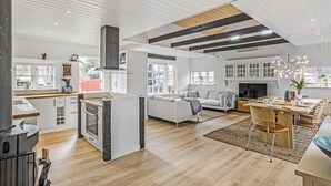 A spacious living area with a fireplace, a dining area with wooden chairs, and a kitchen with white cabinets and a large stove. - Pet friendly home in Blokhus with WiFi (Blokhus)