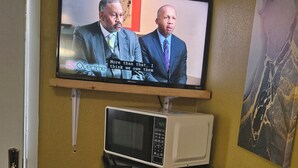 A television mounted on a wall displaying a news broadcast. Below the TV, there is a microwave oven and a coffee maker on a countertop. - Charming lodge in delightful Kelso with AC, WiFi (Kelso)