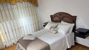Una cama bien hecha con una manta marrón, almohadas blancas y una almohada estampada. Hay dos mesitas de noche con superficie negra y un cabecero de madera. La habitación tiene suelo de madera y cortinas blancas con un estampado.