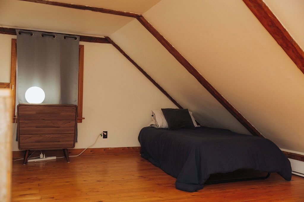 A bedroom with a sloped ceiling, a wooden dresser, a bed with a dark bedspread, and a wall-mounted light fixture.