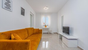 A modern living room with a mustard yellow sofa, a flat-screen TV on a white stand, and a small white table with a vase of flowers. - Amazing apartment in Novalja with WiFi (Novalja)