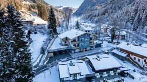 A snow-covered mountain town with a central building featuring multiple floors and balconies, surrounded by snow-laden trees and mountains. - Appartement MaLo - Top 3 (Viehhofen)