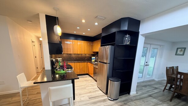 A modern kitchen with wooden cabinets, a stainless steel refrigerator, and a black shelving unit. - Charming 3‑BR Sarasota Retreat — Minutes to Beaches & Downtown (Sarasota)