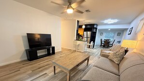 A living room with a flat-screen TV, a wooden coffee table, a sofa with patterned cushions, a dining area with chairs, and a kitchen with modern appliances. - Charming 3‑BR Sarasota Retreat — Minutes to Beaches & Downtown (Sarasota)