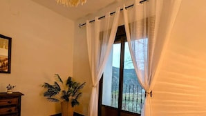 Une chambre avec un grand lit, un luminaire suspendu, une plante en pot et une vue sur un balcon à travers des rideaux transparents.