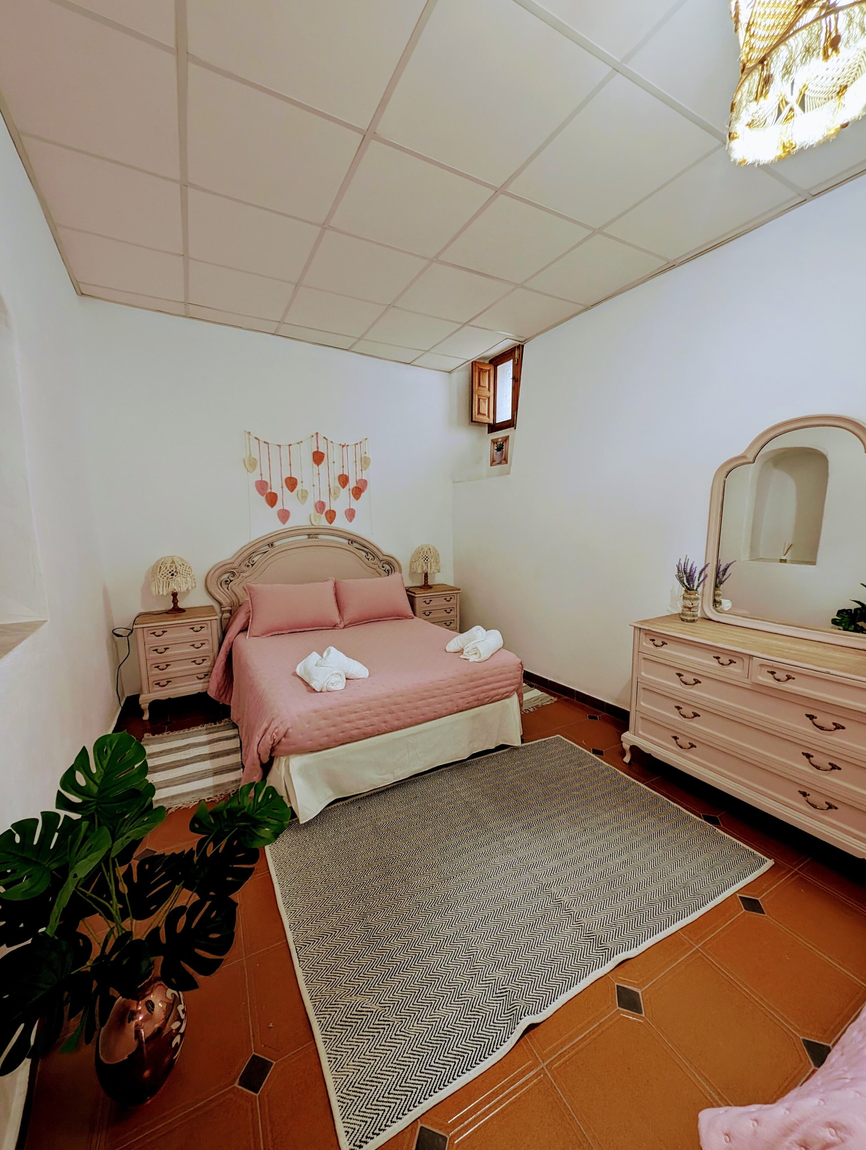 A bedroom with a bed, bedside tables, a dresser, a mirror, a hanging heart decoration, and a potted plant.