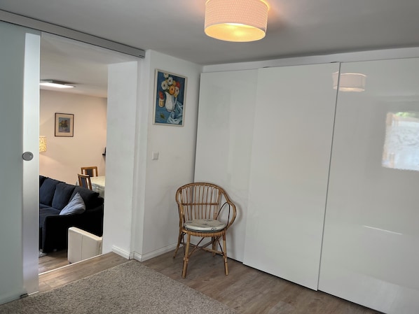 Een kamer met een witte garderobe, een rieten stoel, een klein vloerkleed en een schilderij aan de muur.
