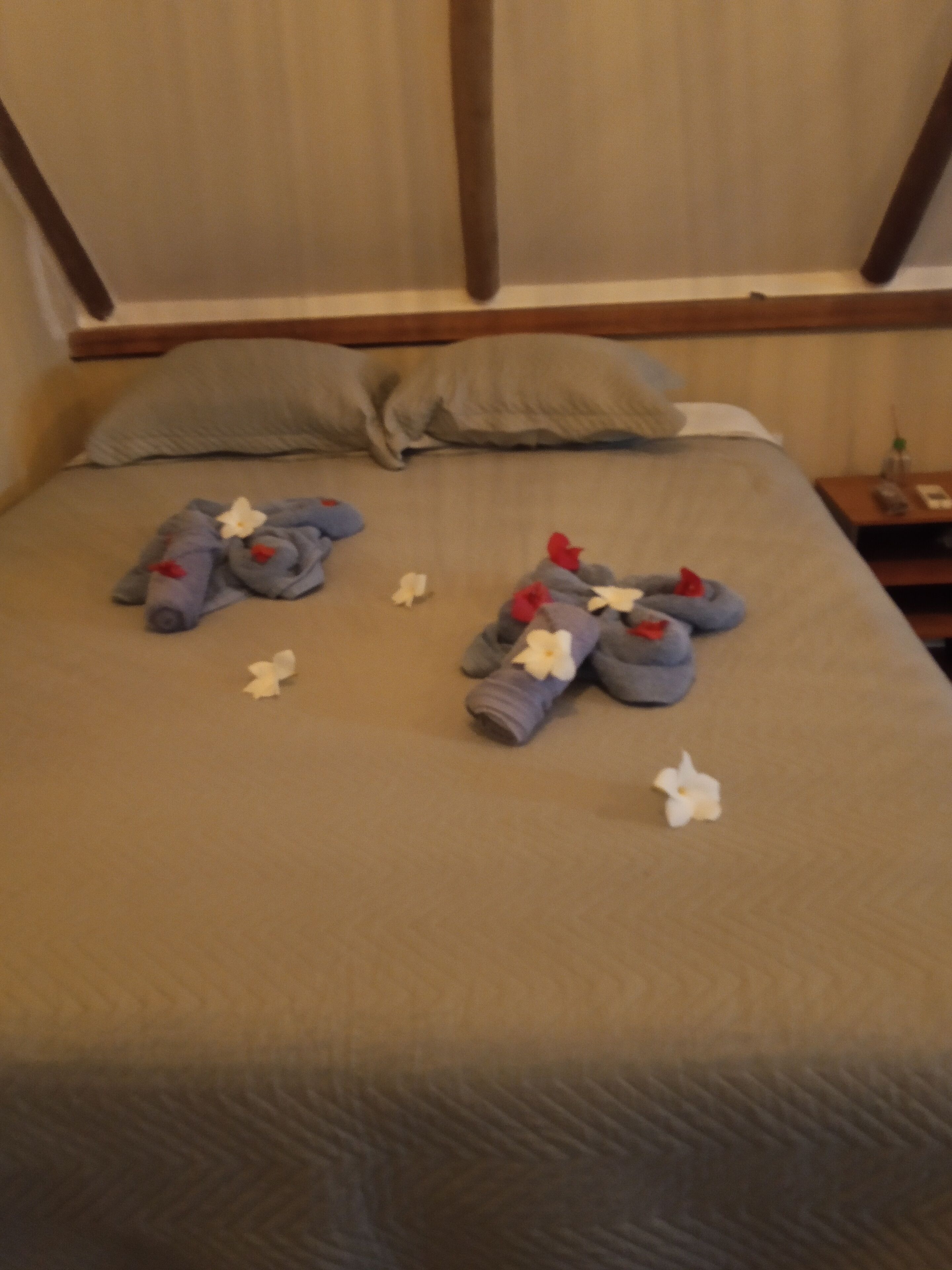 Una cama bien hecha con toallas y flores, un cabecero de madera y una mesita de noche con una botella y un teléfono.