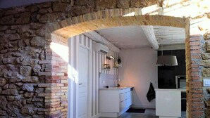 Une cuisine étroite avec des meubles blancs, un mur en pierre d’un côté et un luminaire suspendu.