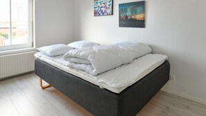 Ein modernes Bett mit grauem Gestell und weißer Bettwäsche in einem Zimmer mit hellem Holzboden und einem großen Fenster.