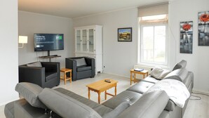 Ein modernes Wohnzimmer mit einer grauen Sofagarnitur, einem Fernseher, einer Kommode und einem Fenster mit Kunstwerk.