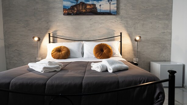 A neatly made bed with a dark brown headboard, two round pillows, and folded white towels. There is a framed picture of a historic building on the wall above the bed. - San Felice Palace (Naples)