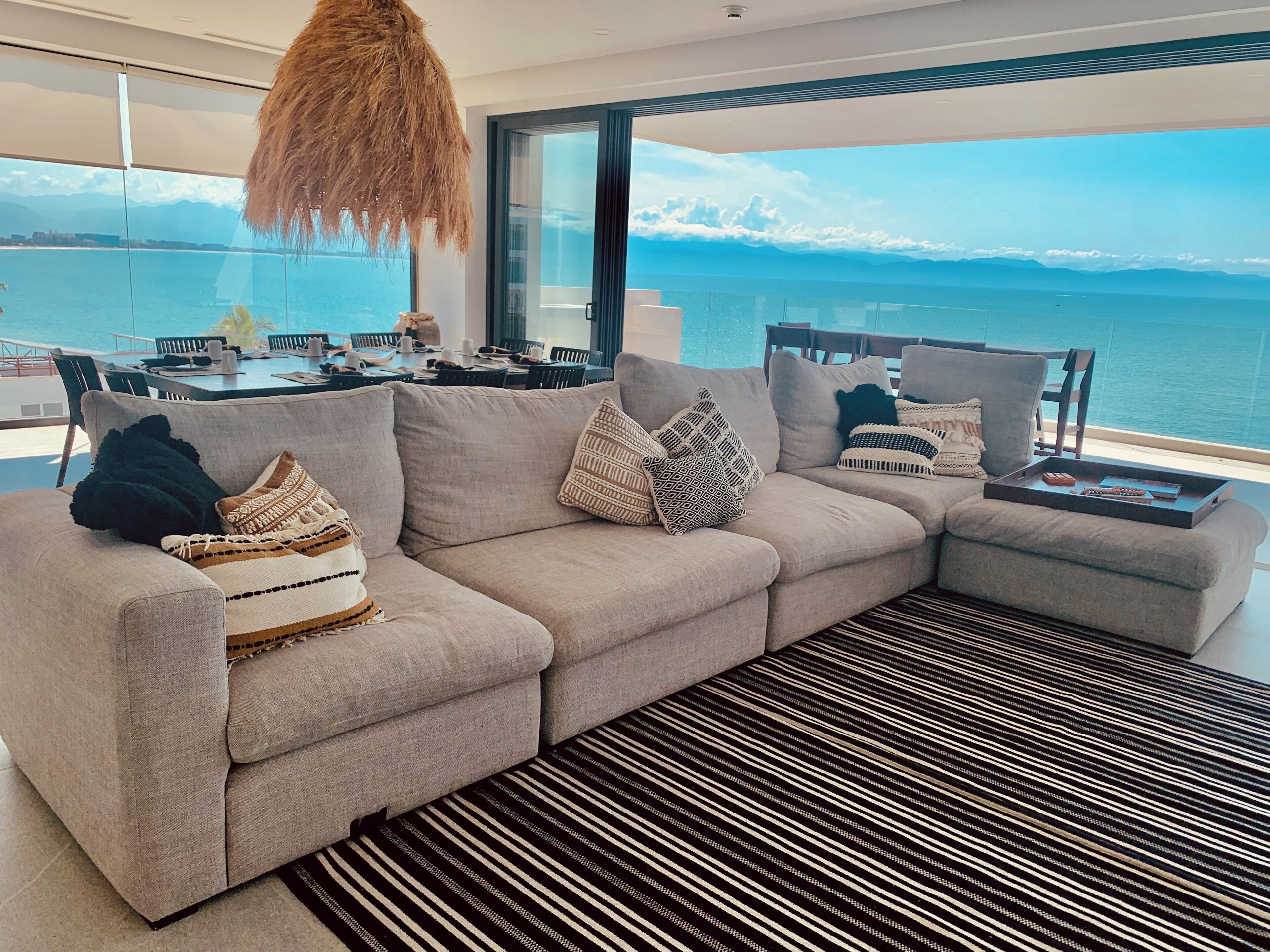 A modern living room with a large sofa, a dining area, and a view of the sea.