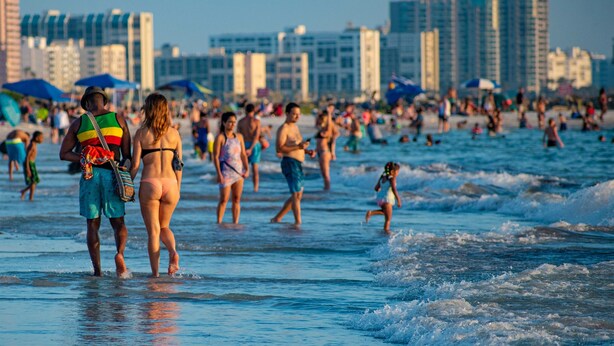 A crowded beach with people walking and swimming, and tall buildings in the background.