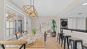 A dining area with a wooden table set for four, surrounded by chairs. A hanging light fixture is above the table, and a 'welcome' sign is on the wall.
