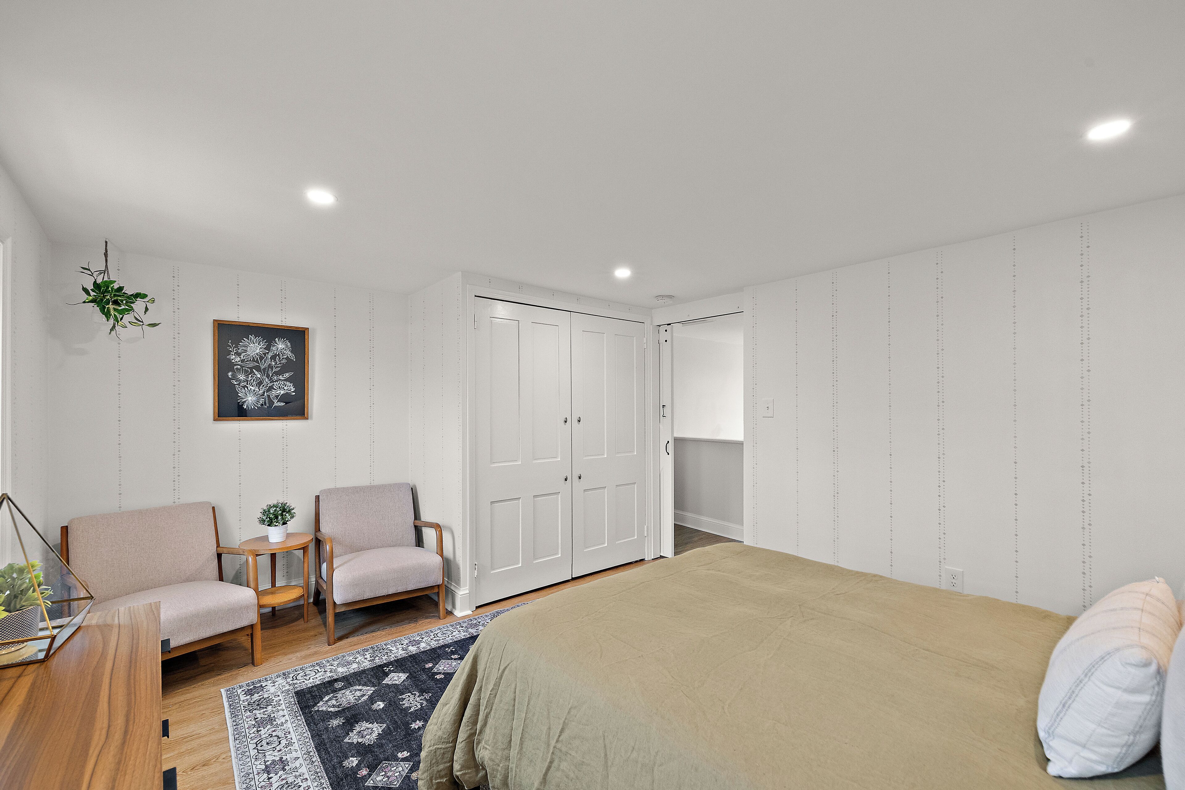 A bedroom with a bed, two chairs, a small table, a plant, and a wardrobe.