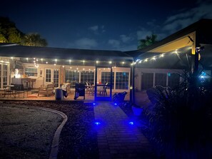 A covered outdoor patio with string lights, a brick pathway, and a seating area with tables and chairs.