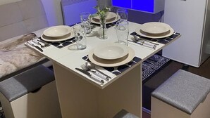 Een moderne eetruimte met een witte tafel gedekt voor vier personen, een flatscreen televisie erboven en een koffieapparaat aan de rechterkant.