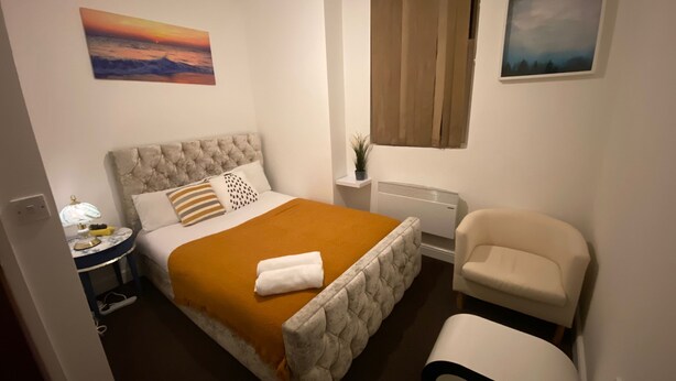 Een slaapkamer met een bed, een stoel, een nachtkastje, een lamp, een radiator en twee schilderijen aan de muur.