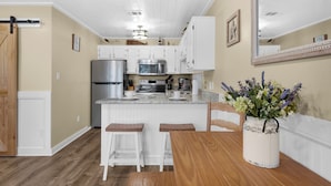 A kitchen with white cabinets, stainless steel appliances, a wooden dining table with two stools, and a vase of flowers.