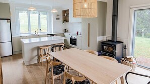 Una cocina moderna con armarios blancos, una isla central, una mesa de comedor con sillas, un ventanal grande y un estufa de leña.