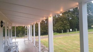 Un porche cubierto con columnas blancas y luces colgantes, una escalera y un banco.