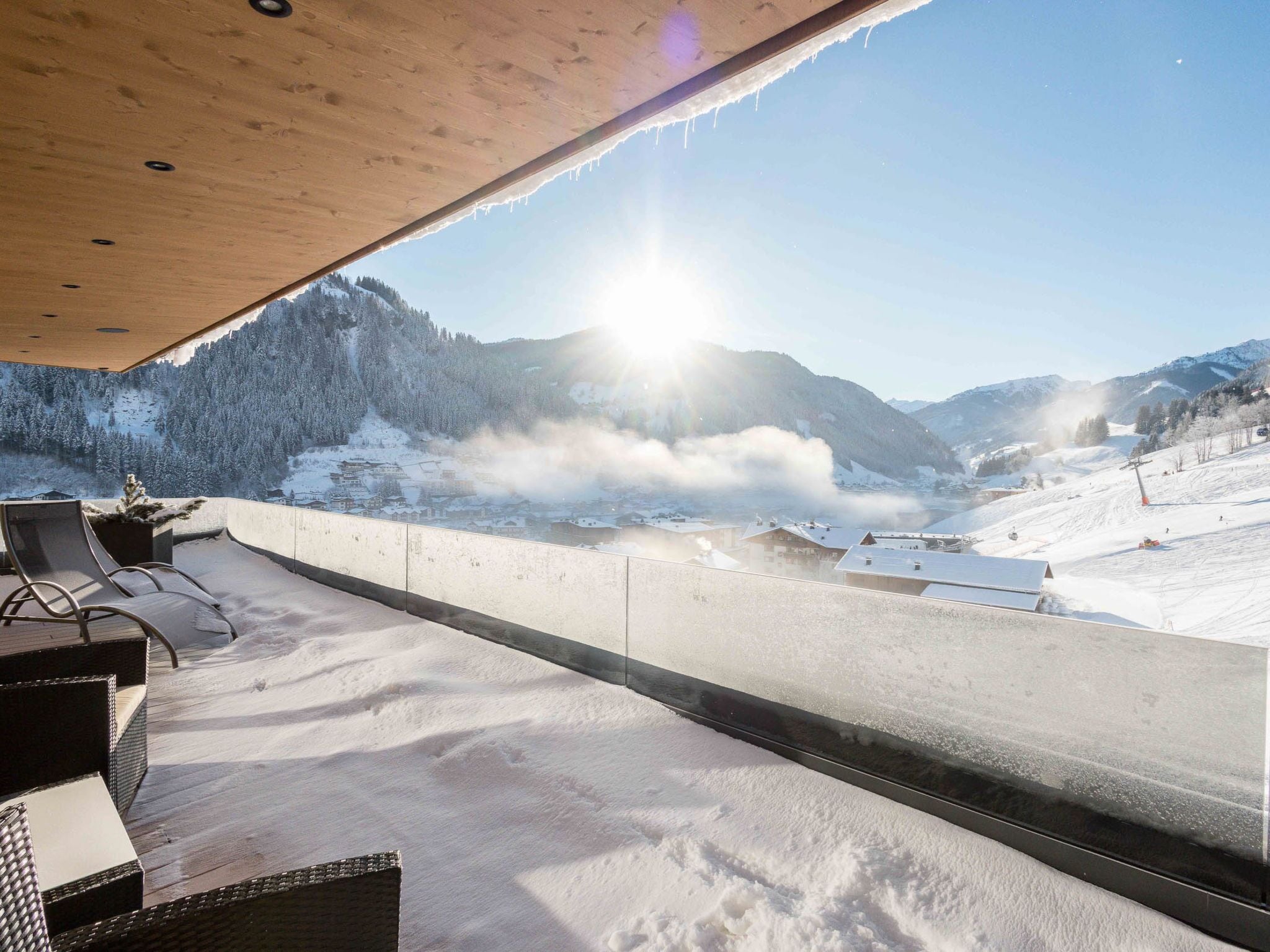 A snow-covered balcony with wicker furniture, overlooking a snowy mountain landscape and a ski slope.