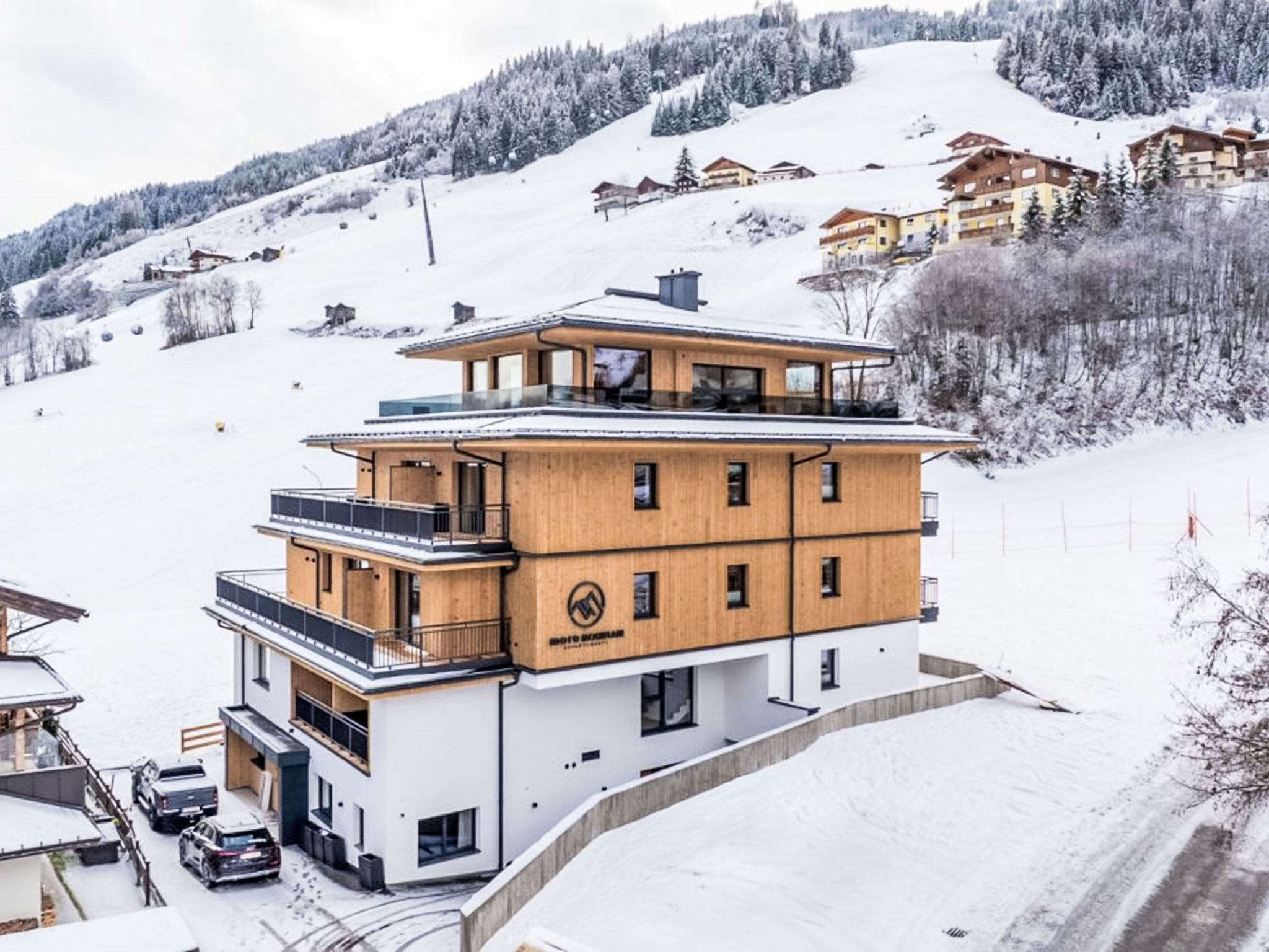 A modern multi-story hotel with a wooden facade, located on a snowy slope with views of surrounding buildings and trees.