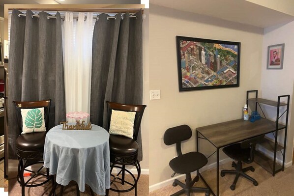 Une pièce comprenant une table à manger, deux chaises, une petite table ronde recouverte d’une nappe, un bureau avec deux chaises et, au mur, un grand tableau encadré représentant un paysage urbain.