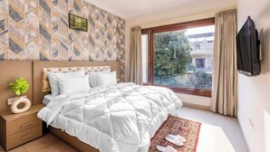 A hotel room with a bed, a TV mounted on the wall, a window with curtains, and a patterned wall. - Nature-inspired luxury 1 BHK (Gurugram)