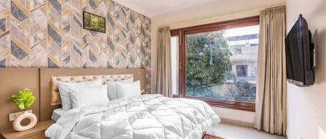A hotel room with a bed, a TV mounted on the wall, a window with curtains, and a patterned wall.