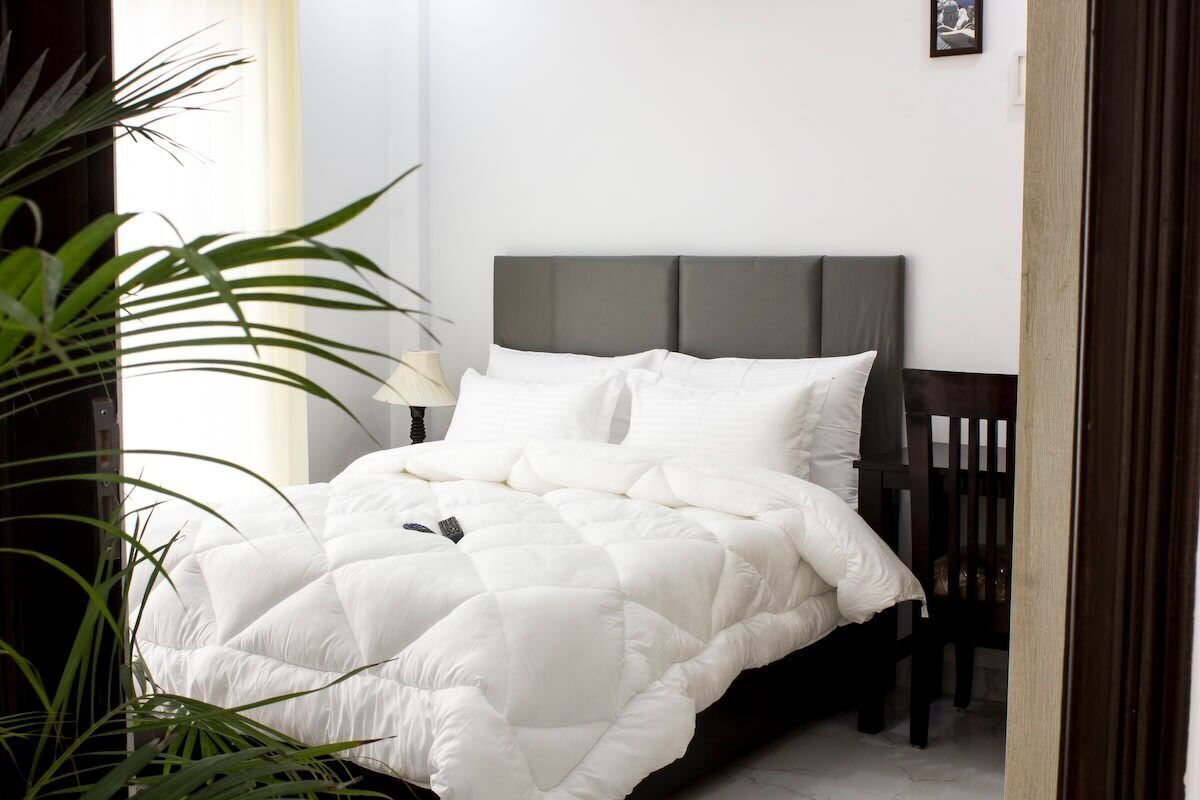 A neatly made bed with a white quilt, a grey headboard, and a bedside table with a lamp.