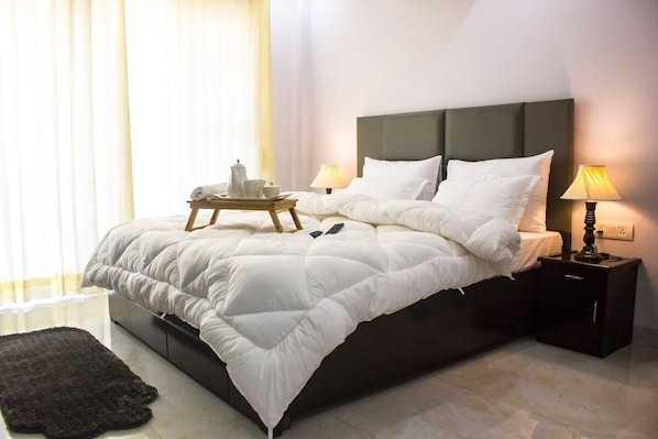 Una cama bien hecha con un cabecero oscuro, una mesita de noche con una lámpara y una pequeña mesa con una bandeja de objetos.