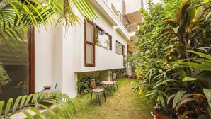 A white building with a small patio area, surrounded by lush greenery and potted plants. - Eco-chic haven in Gurgaon with balcony (Gurugram)