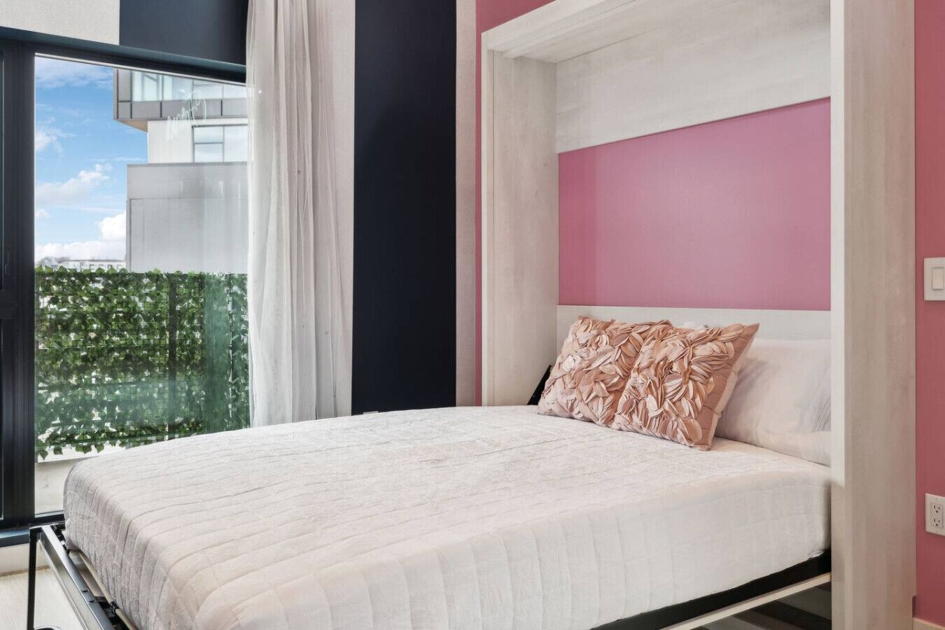 Une chambre moderne avec un grand lit, un mur d’accent rose et une vue sur un immeuble et la verdure environnante.