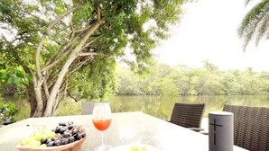 Een tafel met een salade, een fruitschaal en een glas rode drank, voor een achtergrond van een meer en bomen.