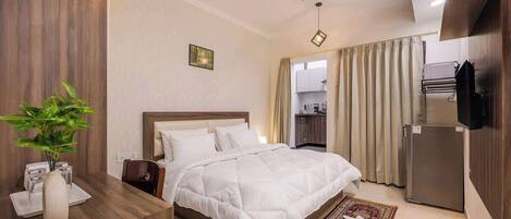 A hotel room with a bed, a desk with a plant, a ceiling fan, and a view of the kitchen.