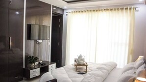 A modern hotel room with a large bed, a TV mounted on the wall, a bedside table with a lamp, and a window with curtains. - Parfait Street 3Bhk Service Apartment Near Fortis (Gurugram)