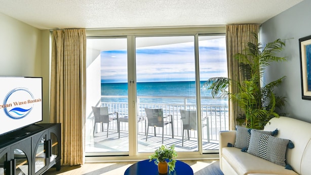 Ein Zimmer mit einer großen Schiebeglastür, die auf einen Balkon mit Meerblick führt. Links befindet sich ein Fernseher, rechts eine Couch mit einer Pflanze und in der Mitte ein blauer Tisch.