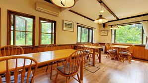 A dining area with wooden tables and chairs, large windows with a view of greenery, and hanging pendant lights. - Pension J-HOUSE (Kiso)