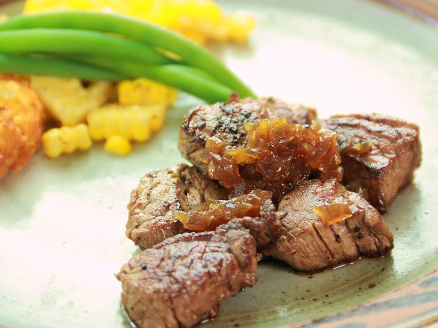 A plate of sliced meat with a caramelized topping, accompanied by yellow corn and green beans.