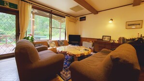 A cozy living room with a wooden ceiling, a large window with curtains, a sofa, a coffee table, and a television. - Pension J-HOUSE (Kiso)