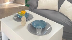 Ein modernes Wohnzimmer mit einem grauen Sofa, weißen Kissen und einem Esstisch, der mit blauen Tellern und einer Vase mit gelben Blumen gedeckt ist.