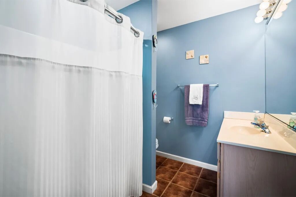 A bathroom with a shower curtain, a towel rack with a purple towel, a sink with a mirror, and a wall-mounted light fixture.