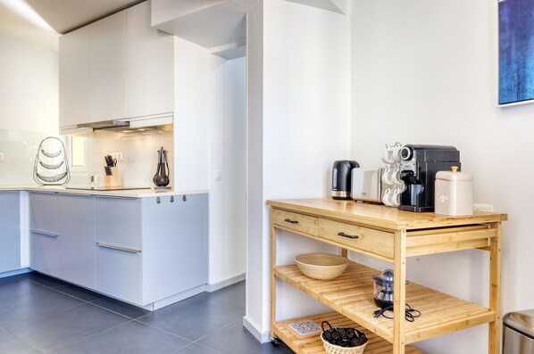 Une cuisine moderne avec des meubles blancs, une table en bois équipée pour préparer du café et un sol carrelé.