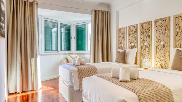 A hotel room with two beds, a window with blinds, a decorative wall panel, and a bedside table with a lamp. - Funchal Panoramic View, a Home in Madeira (Funchal)