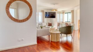 A modern living room with a round mirror, a flat-screen TV, and a dining area with a table and chairs. - Funchal Panoramic View, a Home in Madeira (Funchal)
