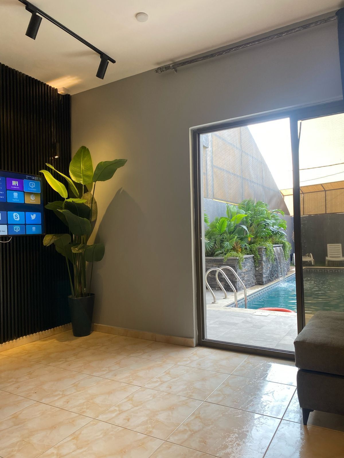 A modern hotel lobby with a large digital screen displaying social media, a potted plant, a sliding glass door to an outdoor pool area, and a seating area.