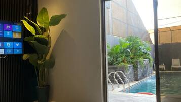 A modern hotel lobby with a large digital screen displaying social media, a potted plant, a sliding glass door to an outdoor pool area, and a seating area.